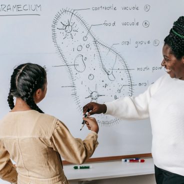 Smiling African American female teacher in casual clothes standing near whiteboard with scheme and helping pupil to write answer